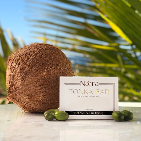 Naera Luxury Soap | Tonka Bar virgin fruit oil bar soap. Handcrafted in the USA with virgin coconut oil and extra virgin olive oil. Picture of soap box on marble table with coconut and olives. Backdrop is palms, the blue sky and the blue ocean. Topics: olive soap, coconut soap, best of nature beauty, tonka soap. body care, face care, scalp care, men's soap, women's soap, gender neutral soap, self care